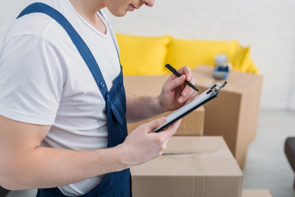 cropped view of mover writing in clipboard near cardboard boxes in apartment
